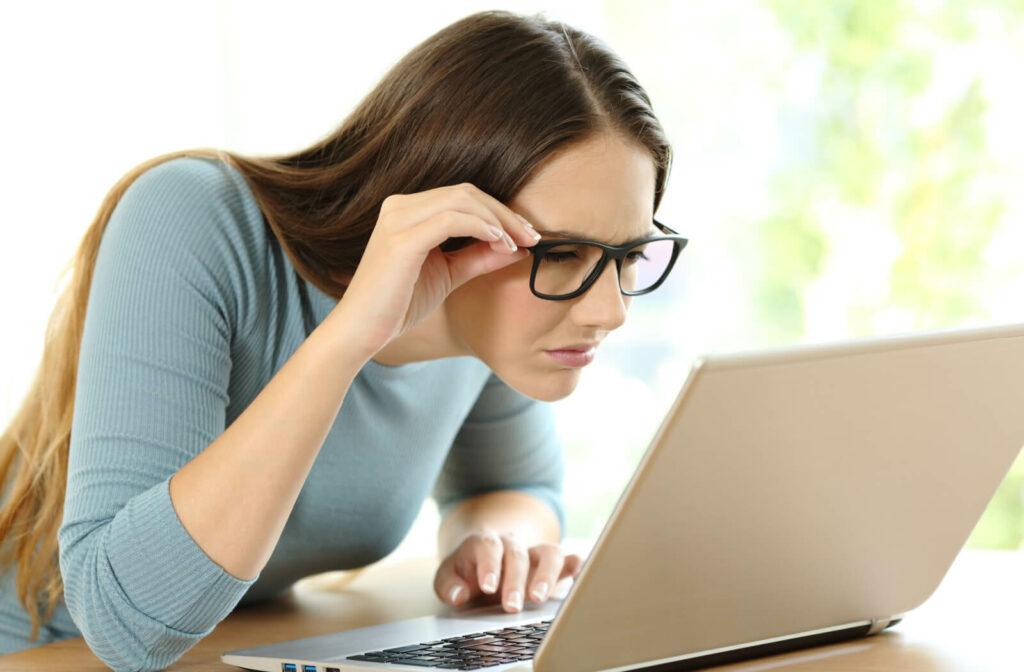 A young woman with myopia having trouble reading, holding her eye glasses ass he moves closer to the laptop to see clearly