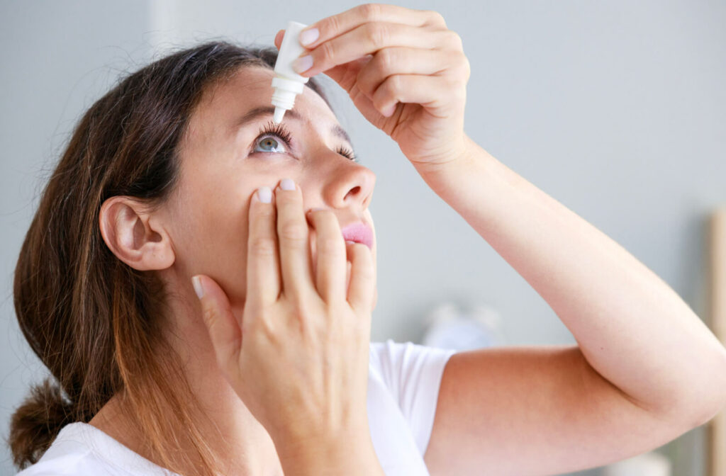 A woman holding a small bottle of eye drops in her left hand and putting them on her right eye while she uses her fingers to pull her eyelid down.